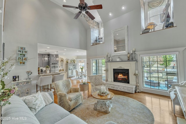 living area featuring a warm lit fireplace, ceiling fan, a high ceiling, light wood-style floors, and recessed lighting