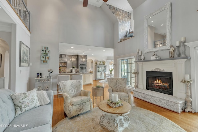 living room featuring a towering ceiling, light wood finished floors, a warm lit fireplace, and arched walkways