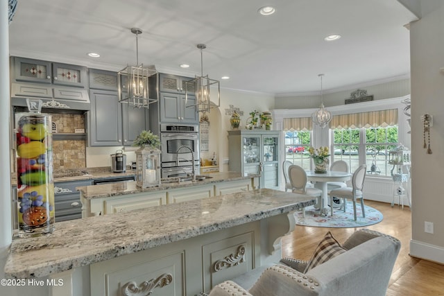 kitchen with light stone counters, stainless steel double oven, gray cabinetry, an island with sink, and crown molding