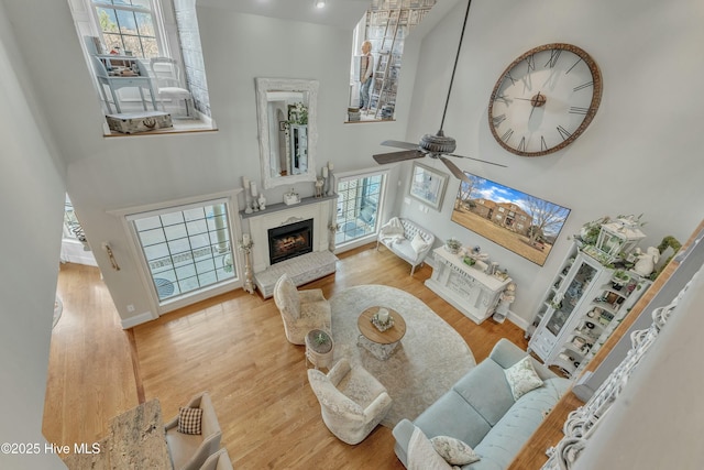 living area with a high ceiling, a ceiling fan, a glass covered fireplace, wood finished floors, and baseboards