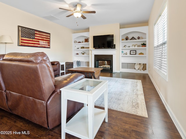 living area with baseboards, built in features, a ceiling fan, a glass covered fireplace, and wood finished floors