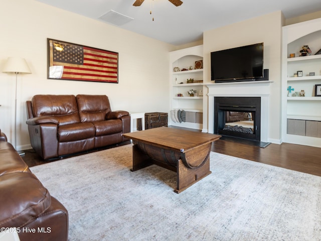 living room featuring built in features, visible vents, a ceiling fan, a glass covered fireplace, and wood finished floors