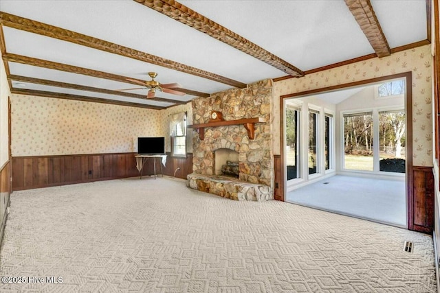 unfurnished living room featuring wallpapered walls, wainscoting, carpet, a stone fireplace, and beam ceiling