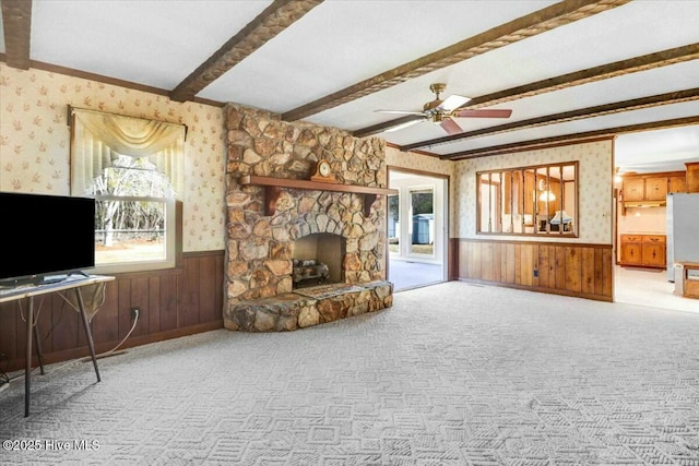 unfurnished living room with carpet, beam ceiling, wainscoting, a stone fireplace, and wallpapered walls
