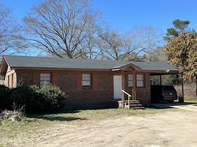 view of front of property featuring entry steps, an attached carport, brick siding, dirt driveway, and roof with shingles