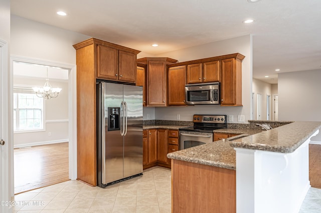 kitchen with brown cabinets, a peninsula, a chandelier, dark stone counters, and stainless steel appliances
