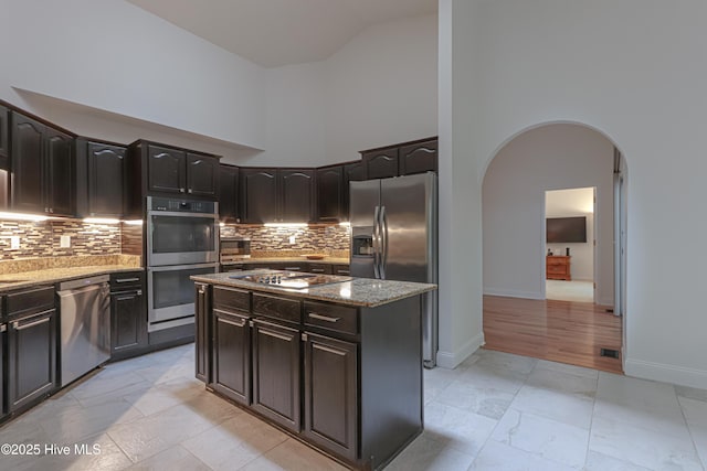 kitchen featuring high vaulted ceiling, light stone counters, backsplash, arched walkways, and appliances with stainless steel finishes