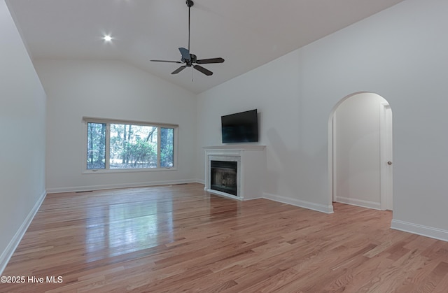 unfurnished living room with high vaulted ceiling, a glass covered fireplace, light wood-type flooring, a ceiling fan, and baseboards