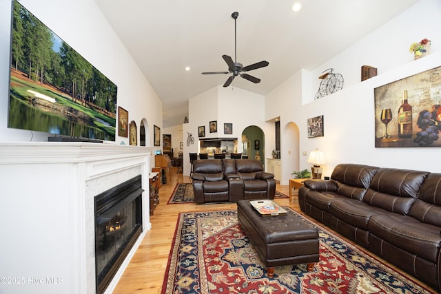 living area featuring a warm lit fireplace, high vaulted ceiling, ceiling fan, arched walkways, and light wood-type flooring