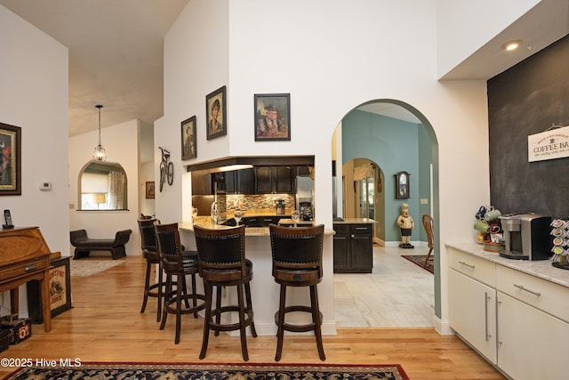 kitchen featuring arched walkways, a towering ceiling, decorative backsplash, light wood-style floors, and a kitchen bar