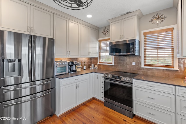 kitchen with a textured ceiling, stainless steel appliances, light wood-type flooring, tasteful backsplash, and dark countertops