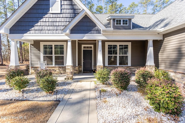 entrance to property featuring covered porch, stone siding, and roof with shingles