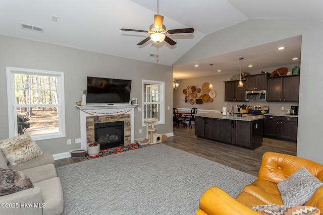 living area with a stone fireplace, ceiling fan with notable chandelier, dark wood-style flooring, visible vents, and vaulted ceiling