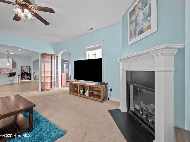 carpeted living room featuring arched walkways, a fireplace with flush hearth, visible vents, and a ceiling fan