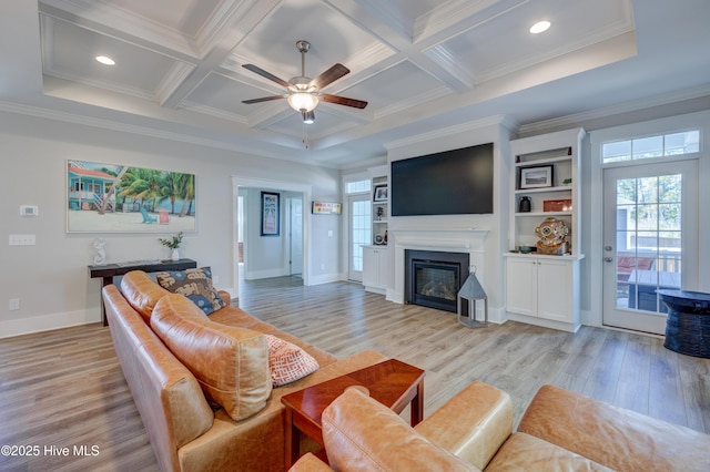 living area featuring light wood finished floors, coffered ceiling, a glass covered fireplace, beamed ceiling, and crown molding