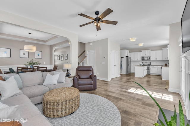 living room featuring light wood-type flooring, arched walkways, baseboards, ceiling fan, and stairs