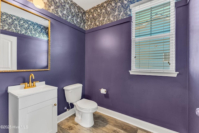 half bath with toilet, wood finished floors, wallpapered walls, baseboards, and vanity