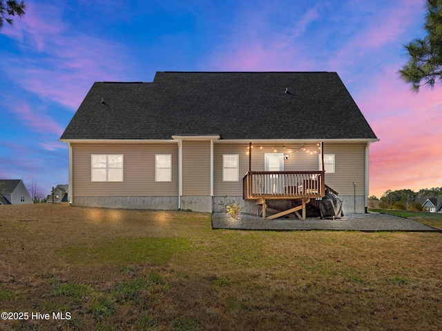 back of property at dusk featuring roof with shingles, a lawn, and a deck