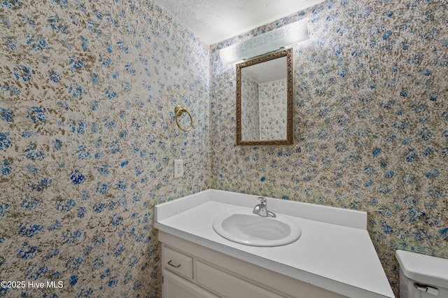 bathroom featuring a textured ceiling, vanity, toilet, and wallpapered walls