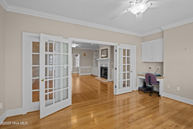 unfurnished office featuring a fireplace with raised hearth, ceiling fan, french doors, ornamental molding, and light wood-type flooring