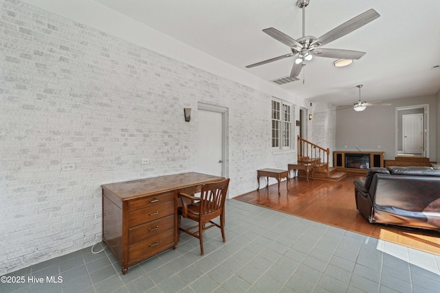 tiled office space featuring brick wall, visible vents, a ceiling fan, and a glass covered fireplace