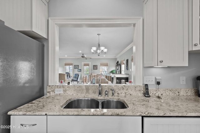 kitchen with light stone counters, ceiling fan with notable chandelier, a sink, white cabinetry, and ornamental molding