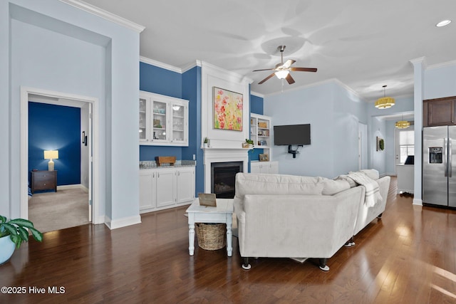 living room featuring dark wood-style flooring, crown molding, a glass covered fireplace, ceiling fan, and baseboards