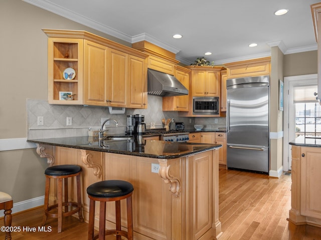 kitchen featuring light wood-style flooring, a sink, built in appliances, a peninsula, and wall chimney exhaust hood