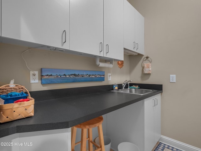 kitchen with a breakfast bar area, dark countertops, white cabinets, a sink, and baseboards