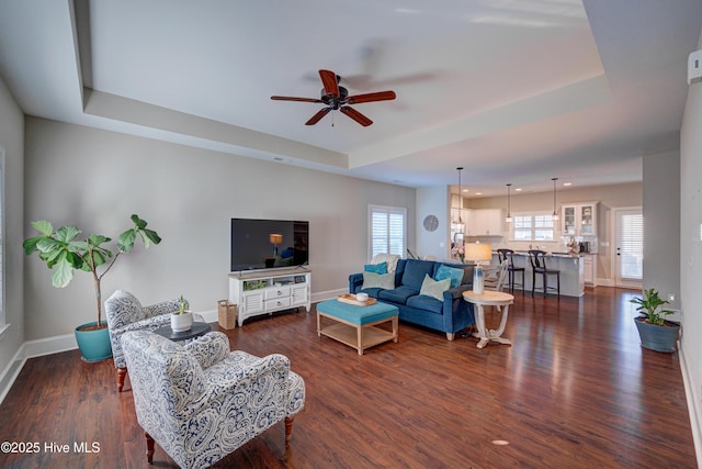 living area featuring a tray ceiling, dark wood-type flooring, a ceiling fan, and baseboards