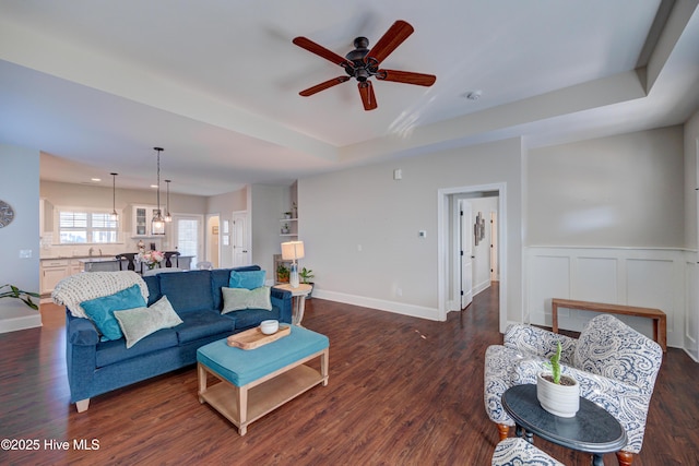 living area with a ceiling fan, a tray ceiling, dark wood finished floors, and baseboards