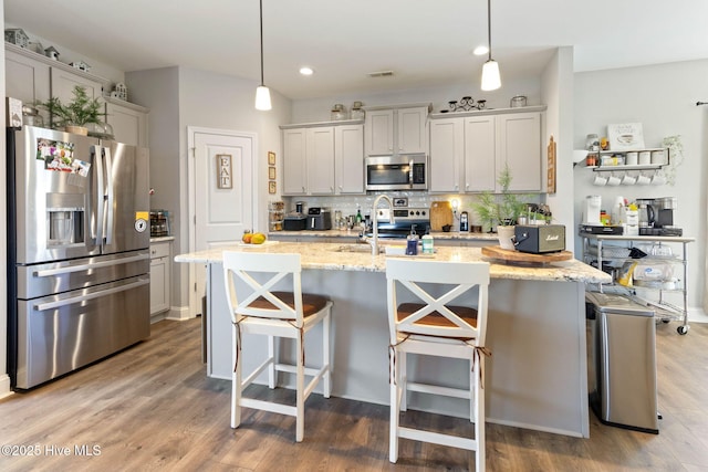 kitchen with a kitchen island with sink, stainless steel appliances, wood finished floors, decorative backsplash, and light stone countertops