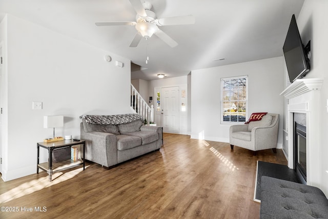 living room featuring a fireplace with flush hearth, a ceiling fan, wood finished floors, baseboards, and stairs