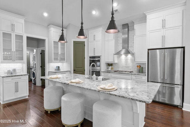2900 Legends Dr, Southport, NC 28461 - kitchen featuring stainless steel appliances, wall chimney range hood, decorative backsplash, glass insert cabinets, and recessed lighting