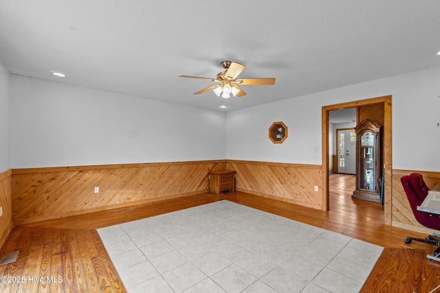 empty room featuring a ceiling fan, a textured ceiling, wood walls, wainscoting, and light wood finished floors