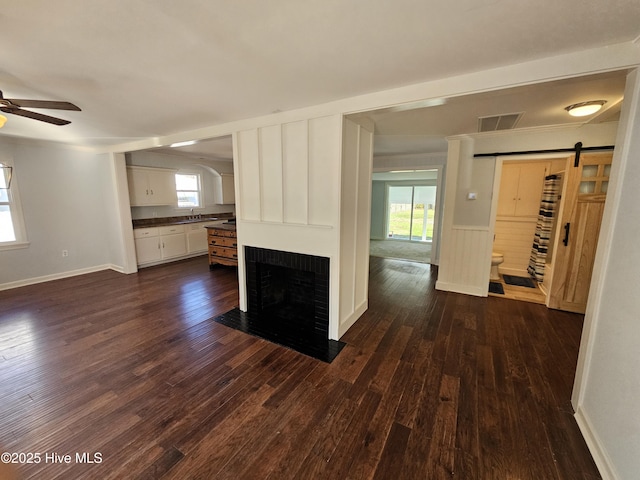 unfurnished living room with visible vents, a barn door, dark wood-type flooring, a ceiling fan, and a brick fireplace