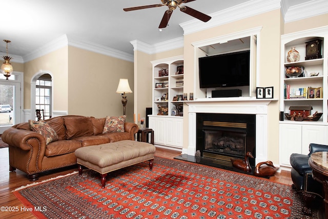 living area featuring a fireplace with raised hearth, ornamental molding, wood finished floors, arched walkways, and a ceiling fan