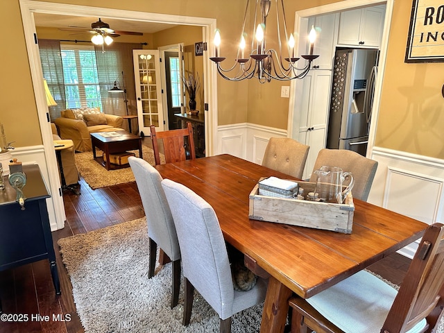 dining area with a wainscoted wall, dark wood-style flooring, ceiling fan with notable chandelier, and a decorative wall