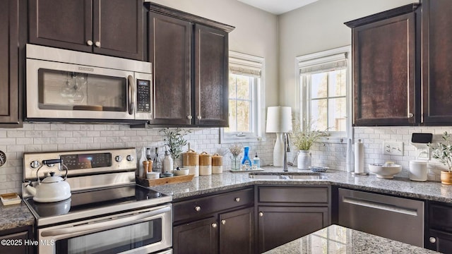 kitchen featuring appliances with stainless steel finishes, a sink, light stone countertops, dark brown cabinets, and backsplash