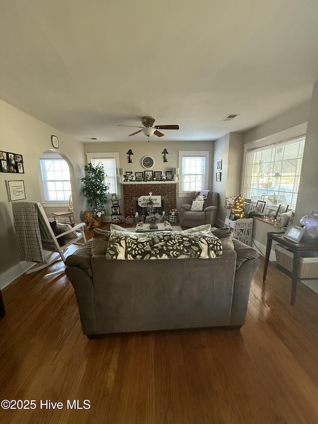 living room with dark wood-style flooring, a ceiling fan, visible vents, baseboards, and a brick fireplace