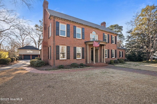 georgian-style home featuring brick siding, an outdoor structure, a detached garage, a chimney, and a front yard