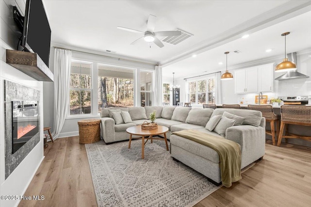 living area featuring visible vents, baseboards, a glass covered fireplace, light wood-style flooring, and ceiling fan