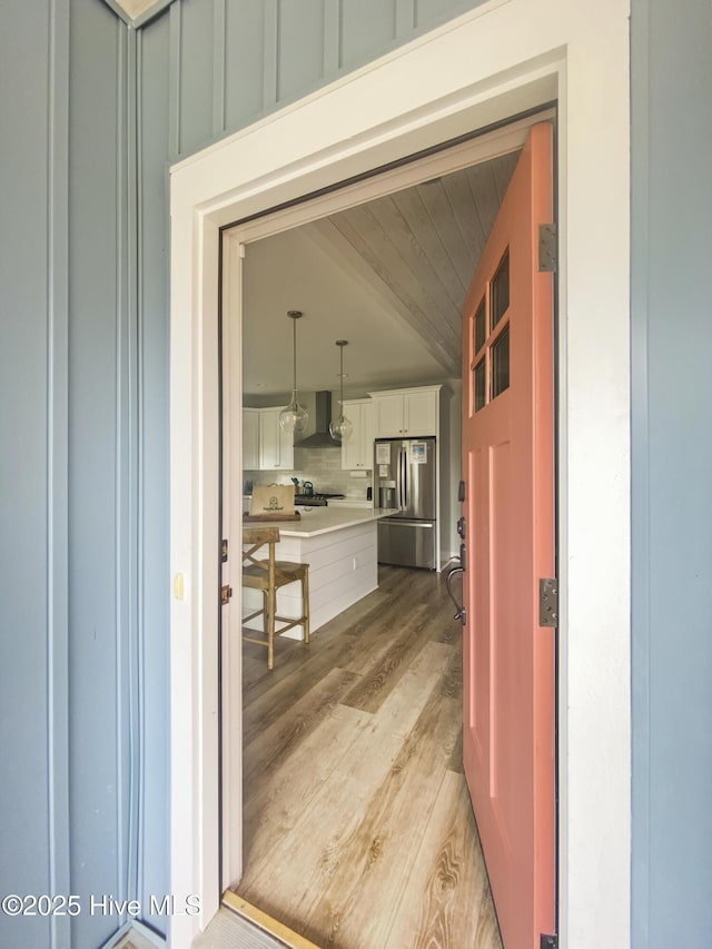 interior space with white cabinets, light wood-style floors, wall chimney range hood, backsplash, and stainless steel fridge with ice dispenser