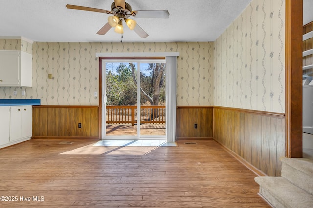 unfurnished dining area with a wainscoted wall, light wood finished floors, ceiling fan, a textured ceiling, and wallpapered walls