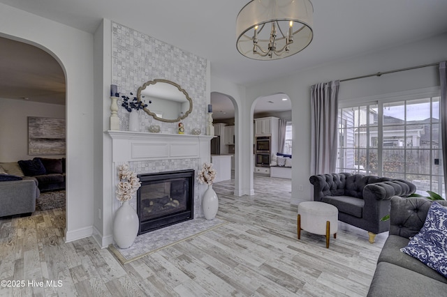 living area featuring arched walkways, a tile fireplace, light wood-style flooring, a notable chandelier, and baseboards