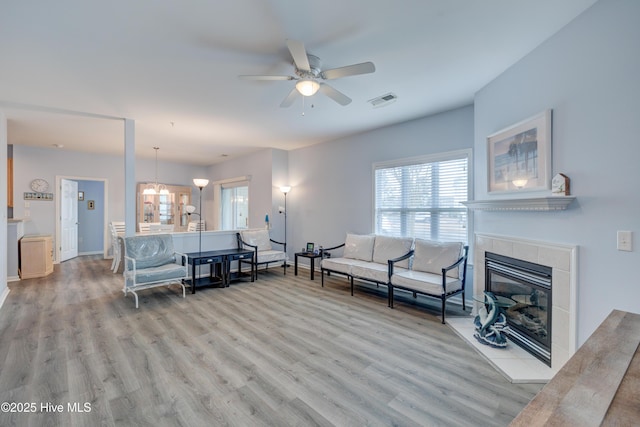 living area featuring a tile fireplace, ceiling fan with notable chandelier, visible vents, baseboards, and light wood-type flooring