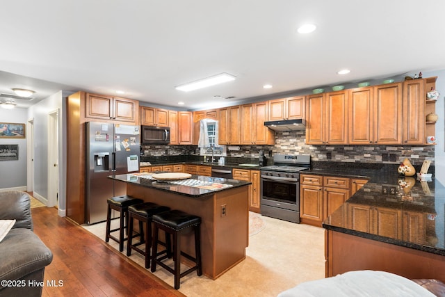 kitchen featuring dark stone countertops, stainless steel appliances, under cabinet range hood, a kitchen bar, and backsplash