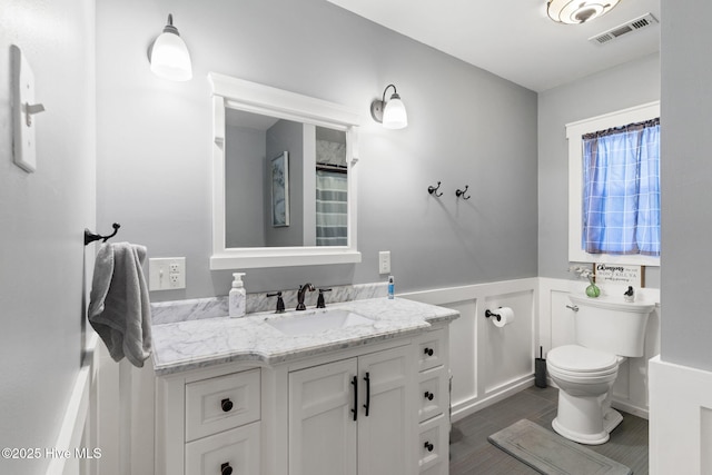 bathroom with visible vents, toilet, a wainscoted wall, vanity, and a decorative wall