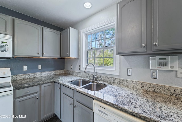 kitchen with light stone counters, white appliances, a sink, and gray cabinetry