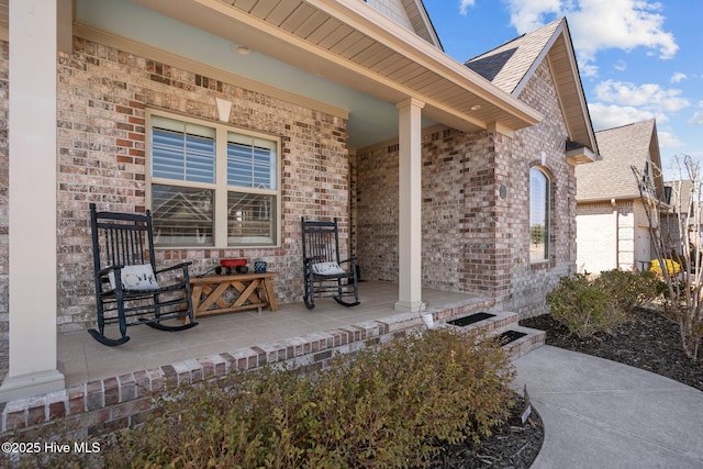 view of exterior entry with brick siding, a porch, and roof with shingles